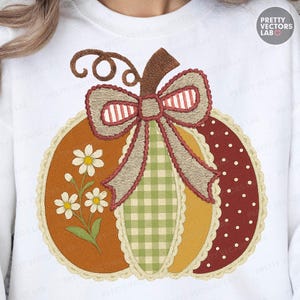 May include: White sweatshirt featuring an embroidered pumpkin design. The pumpkin has sections of orange, yellow, and red with a green gingham and a dotted pattern. A large bow and daisy flowers complete the fall-themed design.