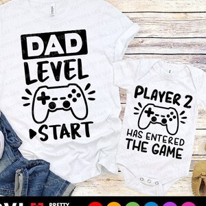 Op de afbeelding: Twee witte T-shirts met zwarte tekst en afbeeldingen. Het grotere shirt zegt "Papa Level" met een afbeelding van een videogamecontroller en een "Start"-pijl. Het kleinere shirt zegt "Speler 2 is het spel ingegaan" met een afbeelding van een videogamecontroller.