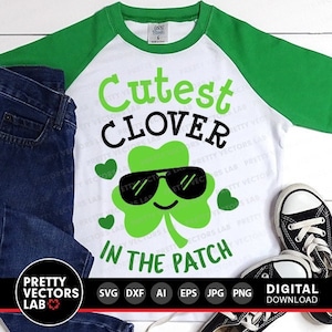 Puede incluir: Una camiseta blanca con mangas verdes y el texto "Cutest Clover in the Patch" en negro. Un trébol de cuatro hojas verde con gafas de sol está en el centro del texto.