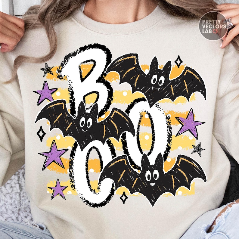 May include: A beige crewneck sweatshirt featuring a graphic print with the word BOO in large white letters outlined in black. Three black cartoon bats with white eyes are positioned around the text, accented by yellow horizontal stripes, white polka dots, and several purple and black stars.