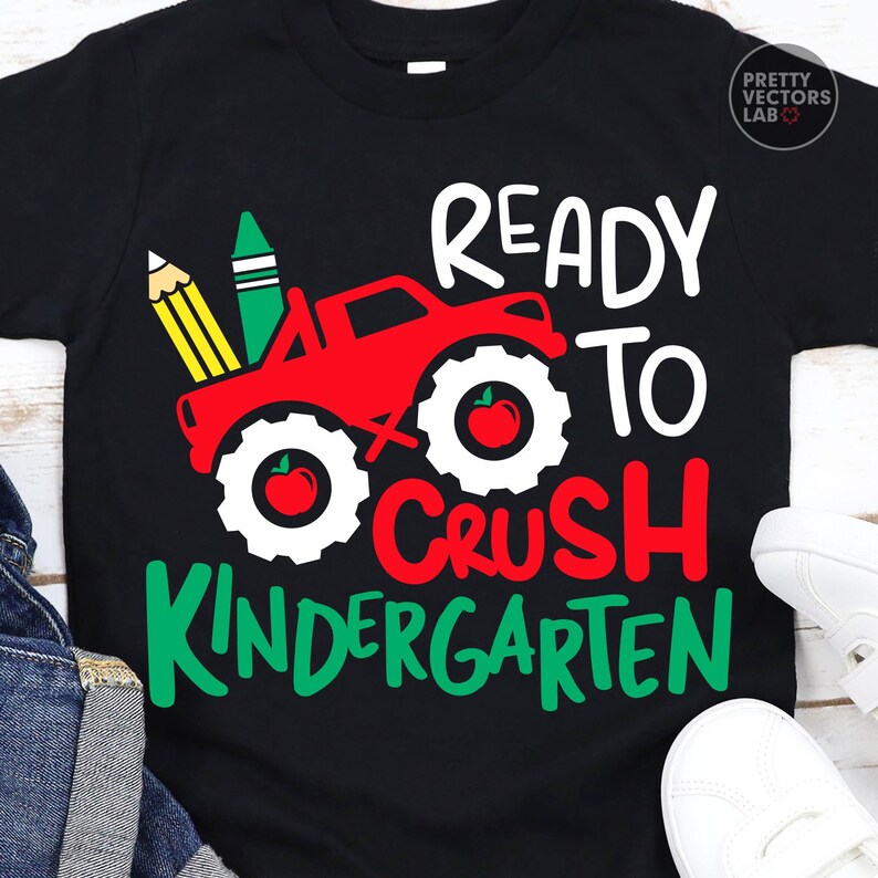Puede incluir: Una camiseta negra con un gr&aacute;fico de un cami&oacute;n monstruo rojo con neum&aacute;ticos blancos, cada uno con una manzana roja. El dise&ntilde;o incluye un l&aacute;piz amarillo, un cray&oacute;n verde y el texto "READY TO CRUSH KINDERGARTEN" en letras blancas y rojas.