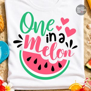 May include: A white t-shirt featuring the text One in a Melon in green, black, and pink script and print fonts. The design includes three pink hearts and a graphic of a watermelon slice with a pink interior, black seeds, and a green rind.