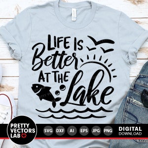 Puede incluir: Una camiseta azul claro con texto negro que dice "Life is Better at the Lake". El texto est&aacute; en una fuente estilizada e incluye un pez saltando fuera del agua y un sol con rayos.