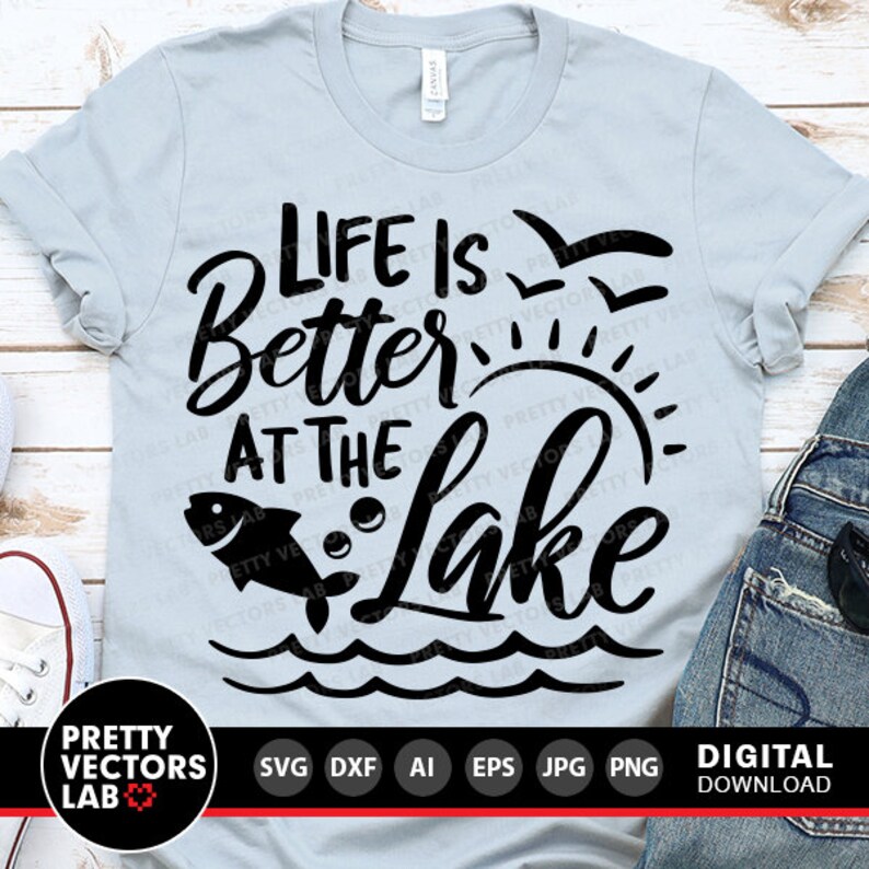 Puede incluir: Una camiseta azul claro con texto negro que dice "Life is Better at the Lake". El texto est&aacute; en una fuente estilizada e incluye un pez saltando fuera del agua y un sol con rayos.