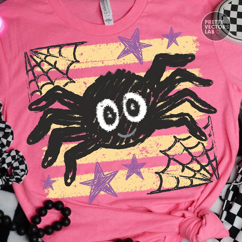 May include: A bright pink t-shirt featuring a central graphic of a black spider with large white eyes, surrounded by purple stars and black spiderwebs over a yellow horizontal striped background. The shirt has a crew neck and a white brand label.