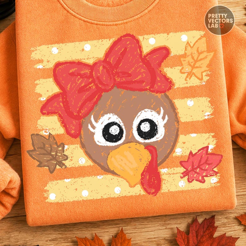 May include: An orange sweatshirt featuring a graphic of a brown turkey face with a large red bow, white eyes with long lashes, and a yellow beak. The background design includes horizontal yellow stripes with white dots and scattered autumn leaves.