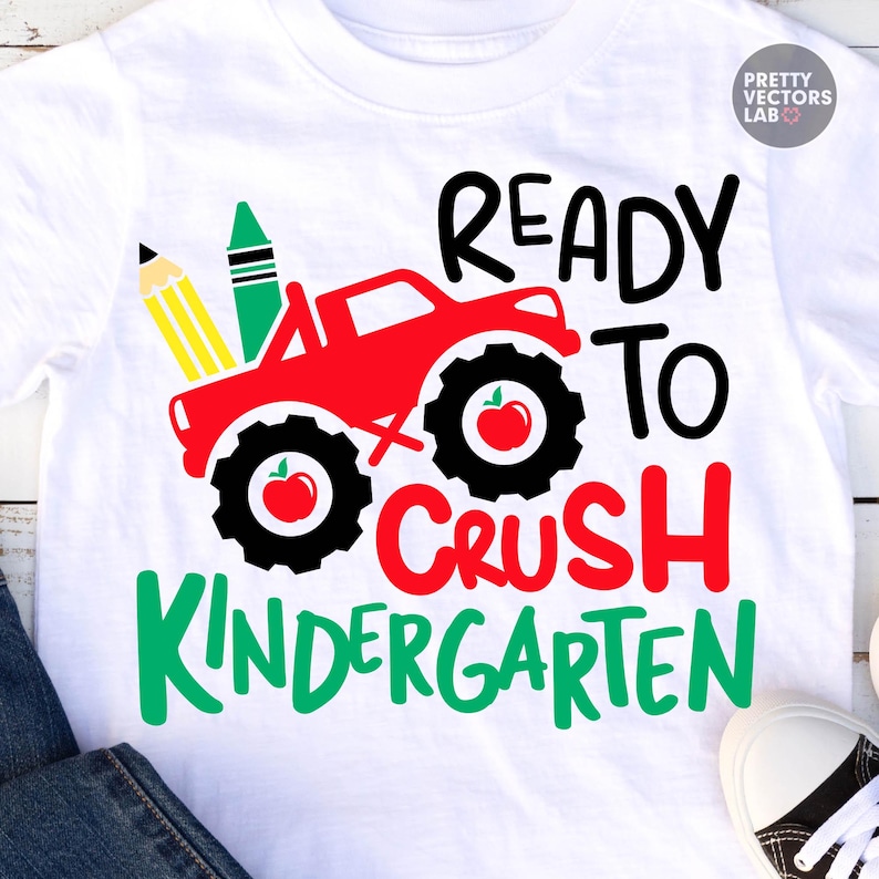 Puede incluir: Una camiseta blanca con un gr&aacute;fico de un cami&oacute;n monstruo rojo con neum&aacute;ticos negros, un l&aacute;piz amarillo y un cray&oacute;n verde. El dise&ntilde;o incluye manzanas rojas dentro de los neum&aacute;ticos y el texto READY TO CRUSH KINDERGARTEN en negro, rojo y verde.