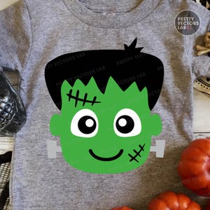 May include: A gray short-sleeved t-shirt featuring a green Frankenstein's monster face graphic. The graphic includes black hair, stitched details, and a smiling face. The shirt is likely for Halloween.