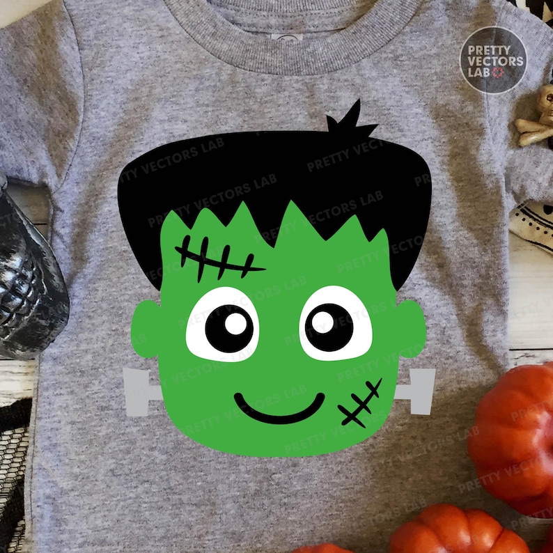May include: A gray short-sleeved t-shirt featuring a green Frankenstein's monster face graphic. The graphic includes black hair, stitched details, and a smiling face. The shirt is likely for Halloween.