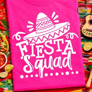 May include: A bright pink t-shirt with the words "Fiesta Squad" in white, along with a white sombrero graphic. The shirt is displayed on a colorful, festive background, suggesting a celebration theme.