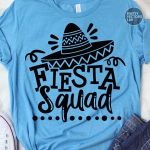 May include: A light blue t-shirt with a black sombrero graphic and the words "FIESTA Squad" in a bold font. The design includes decorative swirls and dots, perfect for a festive occasion.