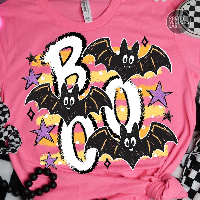 May include: A bright pink t-shirt featuring a graphic print with the word BOO in large white letters. The design includes three black bats with white eyes and small fangs, surrounded by purple stars and yellow horizontal stripes with white dots.