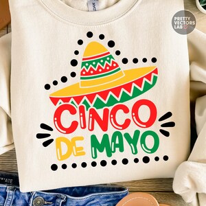 K&ouml;nnte beinhalten: Cremefarbenes Sweatshirt mit einem Cinco de Mayo-Design. Die Grafik zeigt einen gelben Sombrero mit roten und gr&uuml;nen Akzenten und den Text "CINCO DE MAYO" in Rot, Gr&uuml;n und Gelb. Schwarze Punkte und Striche umgeben den Text.