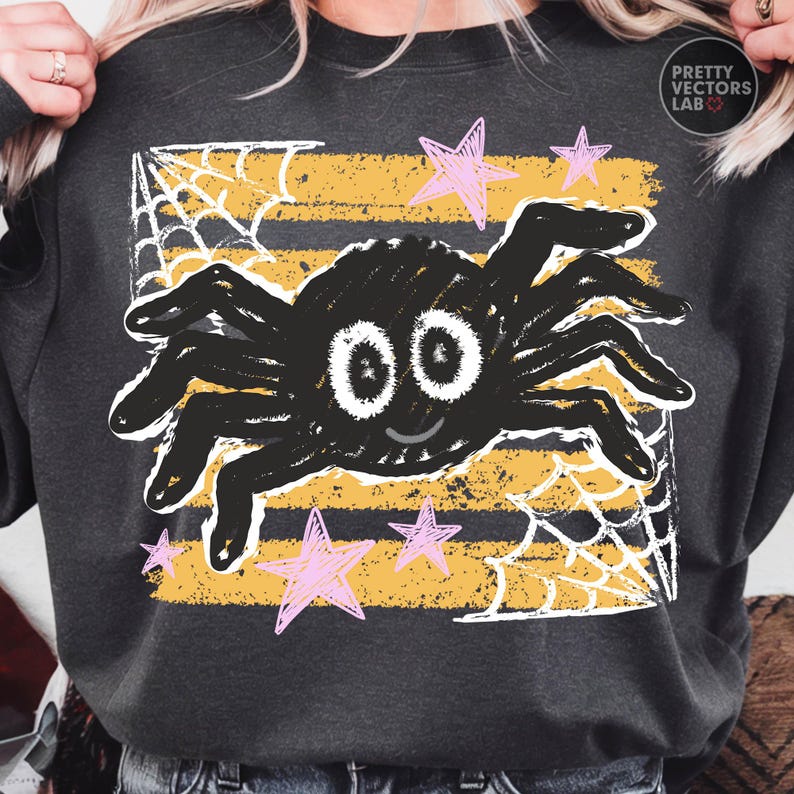 May include: A dark gray sweatshirt featuring a central graphic of a black spider with large white eyes and a small smile. The design includes horizontal yellow distressed stripes, white spiderwebs in the corners, and pink hand-drawn style stars.