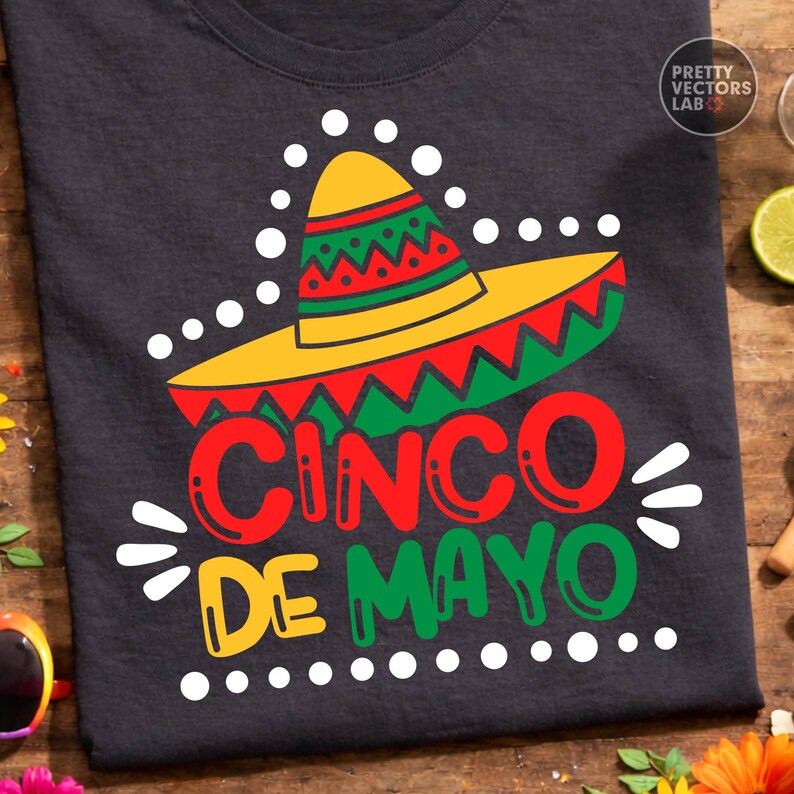 K&ouml;nnte beinhalten: Dunkelgraues T-Shirt mit einem farbenfrohen Cinco de Mayo-Design. Das Design zeigt einen Sombrero in Gelb, Rot und Gr&uuml;n, zusammen mit den Worten "Cinco de Mayo" in fetten, farbigen Buchstaben. Wei&szlig;e Punkte und Akzente vervollst&auml;ndigen den festlichen Look.