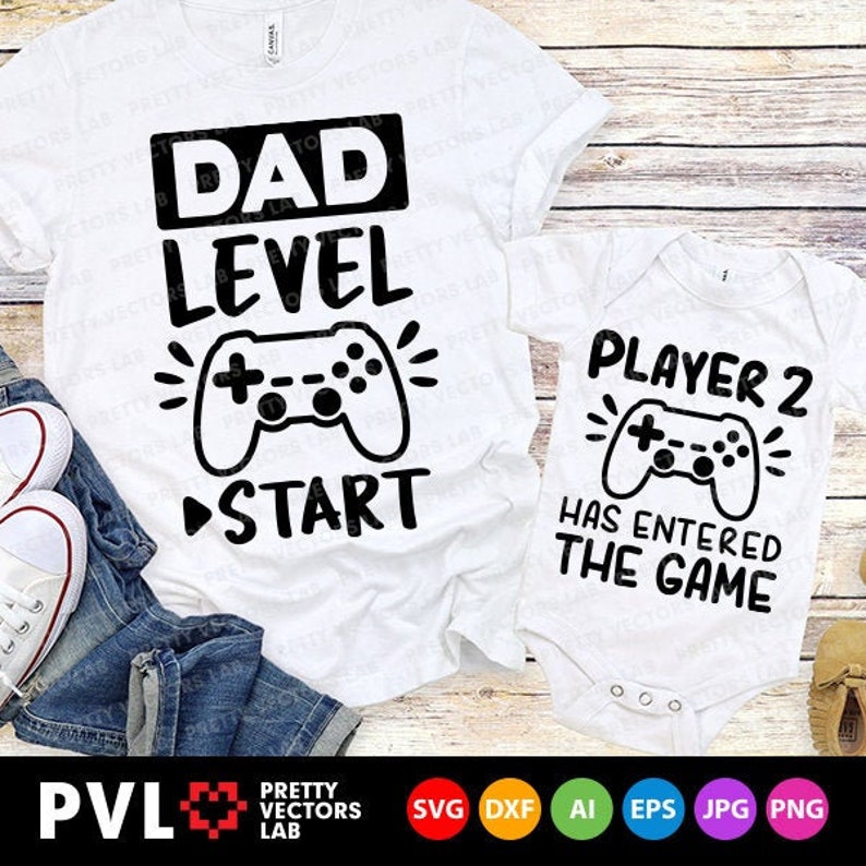 Op de afbeelding: Twee witte T-shirts met zwarte tekst en afbeeldingen. Het grotere shirt zegt "Papa Level" met een afbeelding van een videogamecontroller en een "Start"-pijl. Het kleinere shirt zegt "Speler 2 is het spel ingegaan" met een afbeelding van een videogamecontroller.