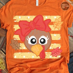 May include: An orange t-shirt featuring a graphic of a turkey face with a large red bow, long eyelashes, and a yellow beak. The design is set against a background of horizontal yellow stripes with white polka dots and scattered autumn leaves.