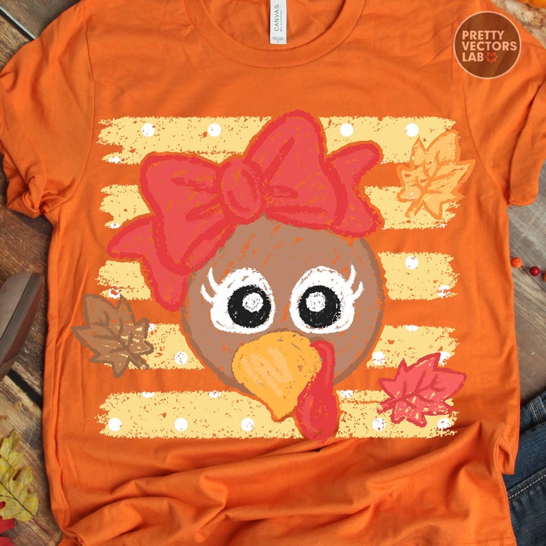 May include: An orange t-shirt featuring a graphic of a turkey face with a large red bow, long eyelashes, and a yellow beak. The design is set against a background of horizontal yellow stripes with white polka dots and scattered autumn leaves.