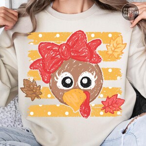 May include: A beige crewneck sweatshirt featuring a graphic of a brown turkey face with large white eyes, long eyelashes, and a bright orange beak. The turkey wears a large red bow. The background has yellow horizontal stripes with white polka dots.