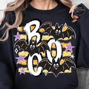 May include: A black long-sleeve sweatshirt featuring a graphic print with the word BOO in large white letters. The design includes three black bats, horizontal yellow and black stripes, and several purple and white stars surrounding the text.