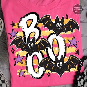 May include: A bright pink t-shirt featuring a graphic print with the text BOO in large white letters. The design includes three black cartoon bats with white eyes and fangs, scattered purple stars, and horizontal yellow stripes with white dots.