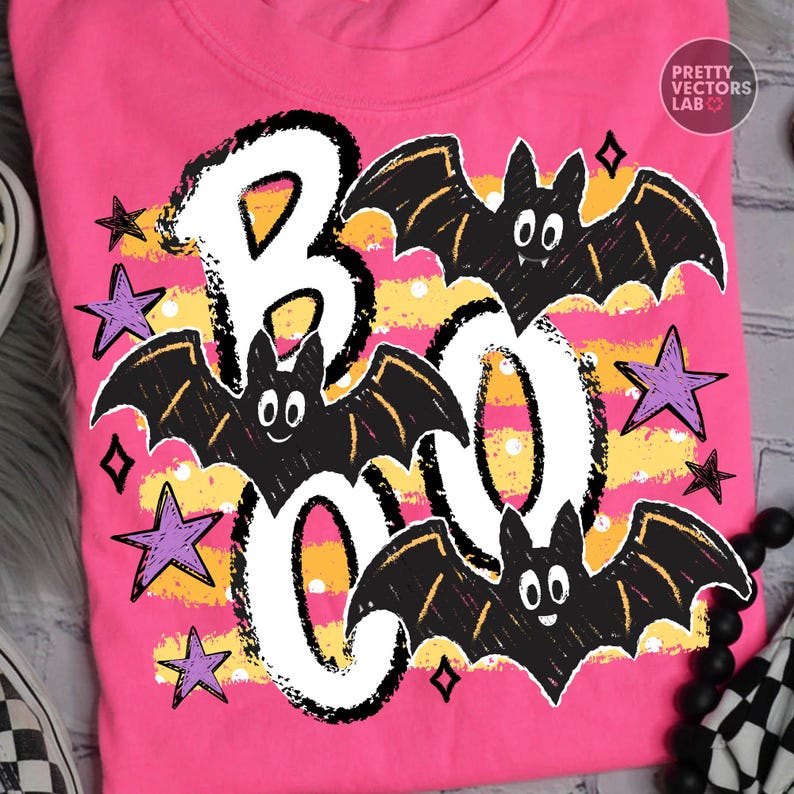 May include: A bright pink t-shirt featuring a graphic print with the text BOO in large white letters. The design includes three black cartoon bats with white eyes and fangs, scattered purple stars, and horizontal yellow stripes with white dots.