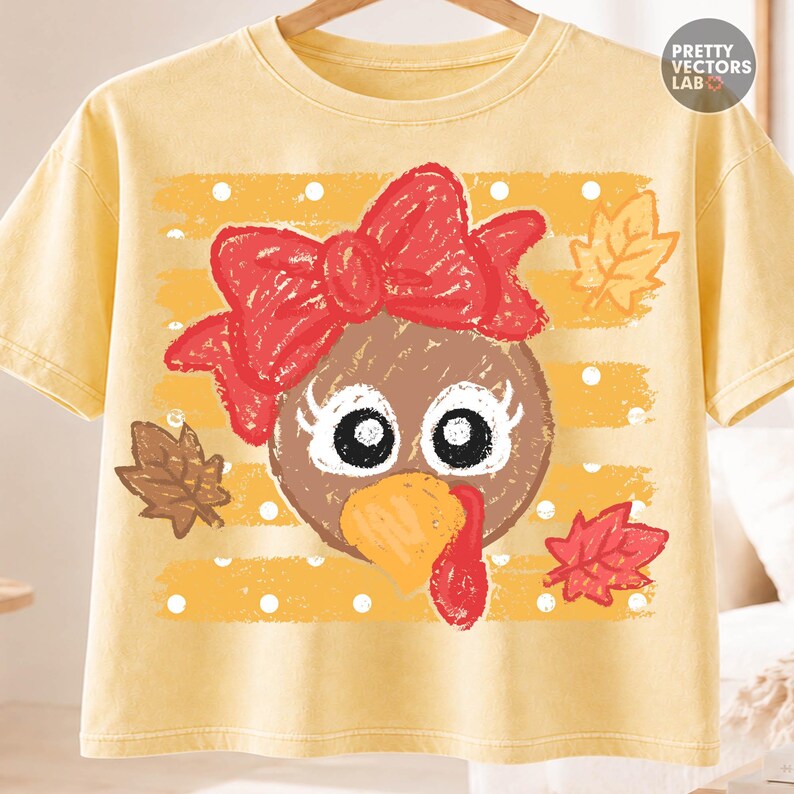 May include: A pale yellow t-shirt featuring a graphic of a brown turkey face with a large red bow, white eyelashes, and an orange beak. The background of the graphic has yellow horizontal stripes with white polka dots and autumn leaves.