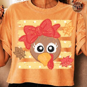 May include: An orange short-sleeve t-shirt featuring a graphic of a turkey face with a large red bow, long eyelashes, and a yellow beak. The design is set against a background of horizontal yellow stripes with white polka dots and autumn leaves.