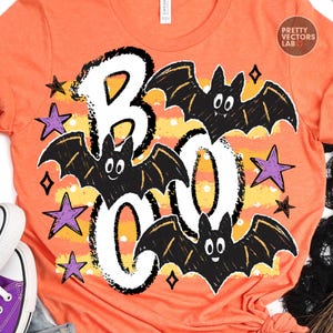 May include: An orange t-shirt featuring a graphic print with the word BOO in large white letters. The design includes three black bats with white eyes and several purple stars. A yellow and orange horizontal striped background with white dots.
