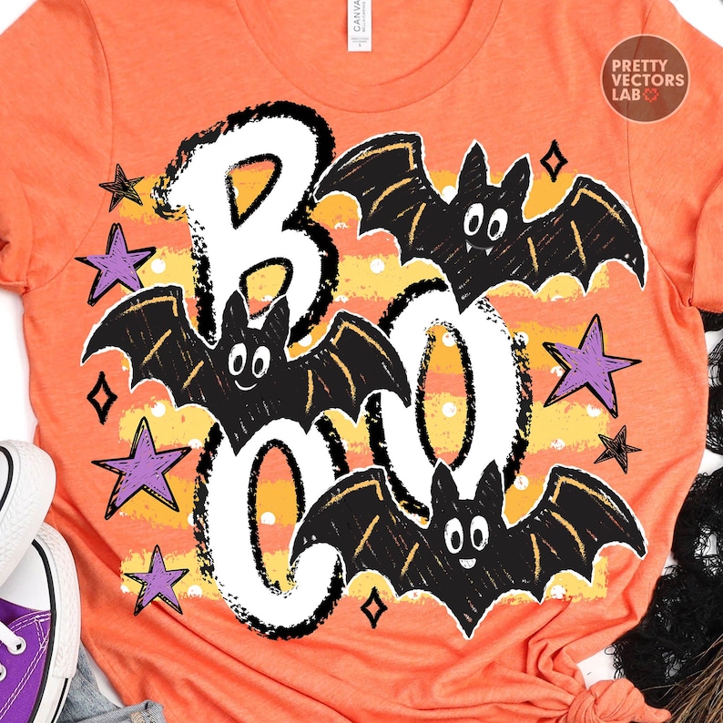 May include: An orange t-shirt featuring a graphic print with the word BOO in large white letters. The design includes three black bats with white eyes and several purple stars. A yellow and orange horizontal striped background with white dots.