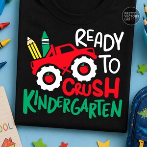 Puede incluir: Una camiseta negra con un gr&aacute;fico de un cami&oacute;n monstruo rojo con neum&aacute;ticos blancos, cada uno con una manzana roja. Sobre el cami&oacute;n hay un l&aacute;piz amarillo y un cray&oacute;n verde. El texto dice READY TO CRUSH KINDERGARTEN en letras blancas, rojas y verdes.