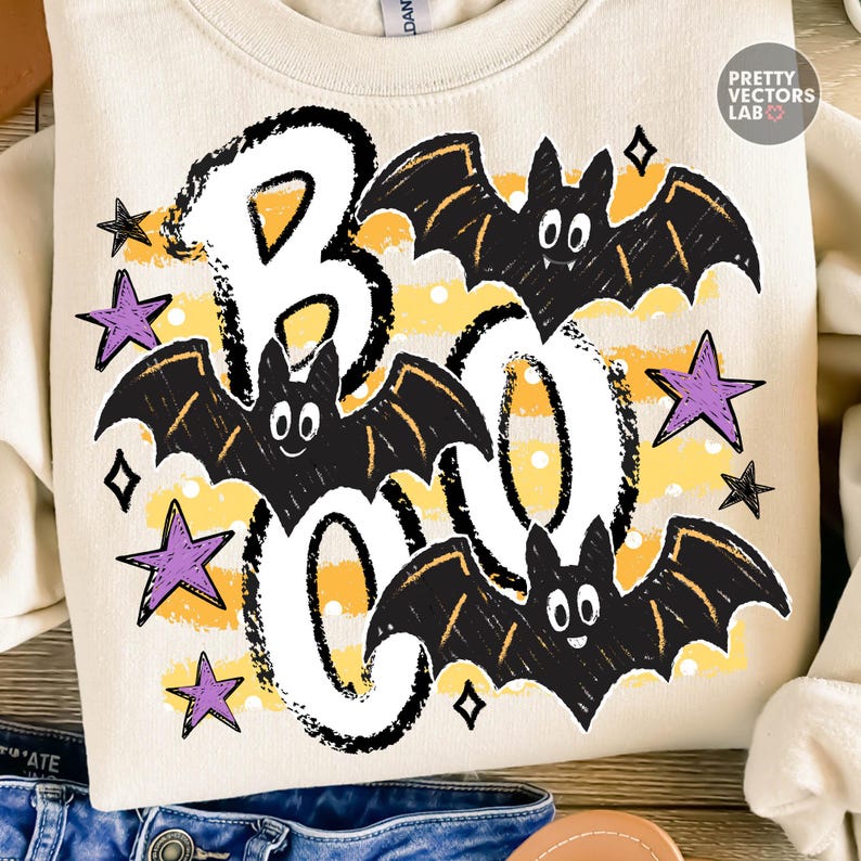 May include: A cream-colored sweatshirt featuring a graphic print with the word BOO in large white letters. The design includes three black bats with white eyes, yellow horizontal stripes, and several purple and black stars scattered throughout.