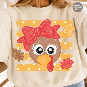 May include: A cream-colored sweatshirt featuring a graphic of a turkey face with a large red bow, long eyelashes, and a yellow beak. The background consists of horizontal yellow stripes with white polka dots and scattered autumn leaves.