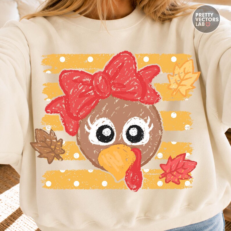 May include: A cream-colored sweatshirt featuring a graphic of a turkey face with a large red bow, long eyelashes, and a yellow beak. The background consists of horizontal yellow stripes with white polka dots and scattered autumn leaves.