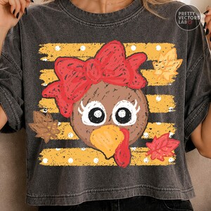 May include: A charcoal gray t-shirt featuring a graphic of a turkey face with a large red bow, long eyelashes, and a yellow beak. The background consists of horizontal yellow stripes with white polka dots and scattered brown and red autumn leaves.
