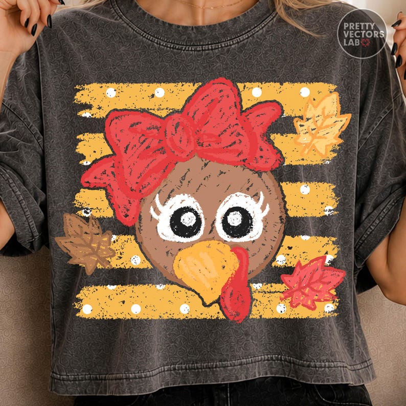 May include: A charcoal gray t-shirt featuring a graphic of a turkey face with a large red bow, long eyelashes, and a yellow beak. The background consists of horizontal yellow stripes with white polka dots and scattered brown and red autumn leaves.