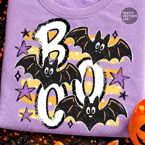May include: A light purple t-shirt featuring a graphic print with the text BOO in large white letters. The design includes three black bats with white eyes, yellow horizontal stripes, and several purple stars scattered across the fabric.