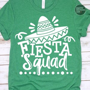 May include: Green t-shirt with a white graphic of a sombrero and the words "Fiesta Squad". The design includes decorative elements and a dotted line below the text. The shirt is made of a soft material.