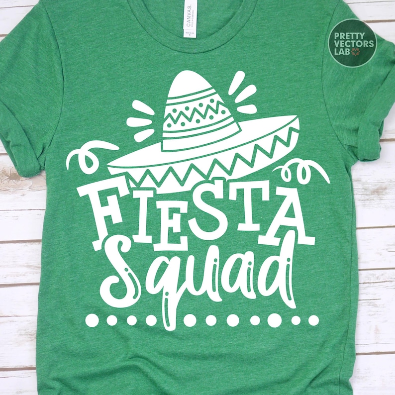 May include: Green t-shirt with a white graphic of a sombrero and the words "Fiesta Squad". The design includes decorative elements and a dotted line below the text. The shirt is made of a soft material.