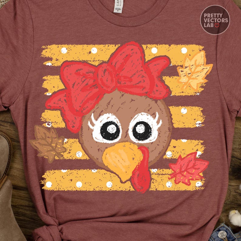 May include: A maroon short-sleeve t-shirt featuring a graphic of a turkey face with a large red bow, white eyelashes, and a yellow beak. The background of the graphic consists of horizontal yellow stripes with white polka dots and autumn leaves.