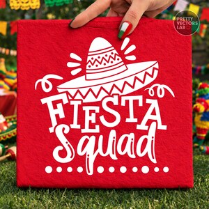 May include: A red square canvas with the words "Fiesta Squad" in white, along with a sombrero graphic. The canvas is held by a hand with painted nails. The background includes festive decorations.