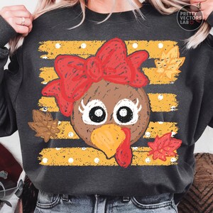 May include: A dark gray sweatshirt featuring a graphic of a brown turkey face with a large red bow, long eyelashes, and a yellow beak. The background consists of horizontal yellow distressed stripes with white polka dots and scattered autumn leaves.