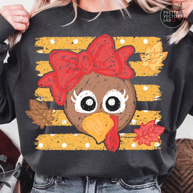 May include: A dark gray sweatshirt featuring a graphic of a brown turkey face with a large red bow, long eyelashes, and a yellow beak. The background consists of horizontal yellow distressed stripes with white polka dots and scattered autumn leaves.