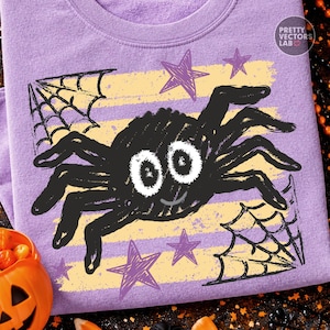 May include: A light purple t-shirt featuring a graphic of a black spider with large white eyes and a small smile. The design includes yellow horizontal stripes, purple stars, and black spiderwebs in the corners. The text Pretty Vectors Lab is visible.