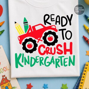Puede incluir: Una camiseta blanca con un gr&aacute;fico de un cami&oacute;n monstruo rojo con neum&aacute;ticos negros, cada uno con un icono de manzana roja. El dise&ntilde;o incluye un l&aacute;piz amarillo, un cray&oacute;n verde y el texto READY TO CRUSH KINDERGARTEN en letras negras y rojas.