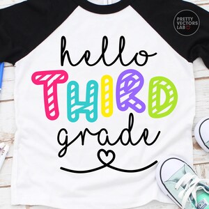 May include: A white raglan t-shirt with black sleeves featuring the black cursive text hello, the colorful striped block text THIRD, and the black cursive text grade with a heart icon below. The design includes pink, teal, yellow, purple, and lime.