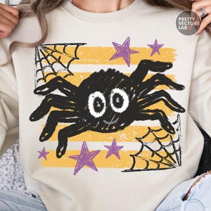 May include: A beige crewneck sweatshirt featuring a graphic of a black spider with large white eyes, surrounded by purple stars and black spiderwebs over horizontal yellow stripes. The text PRETTY VECTORS LAB is printed in the upper right corner.