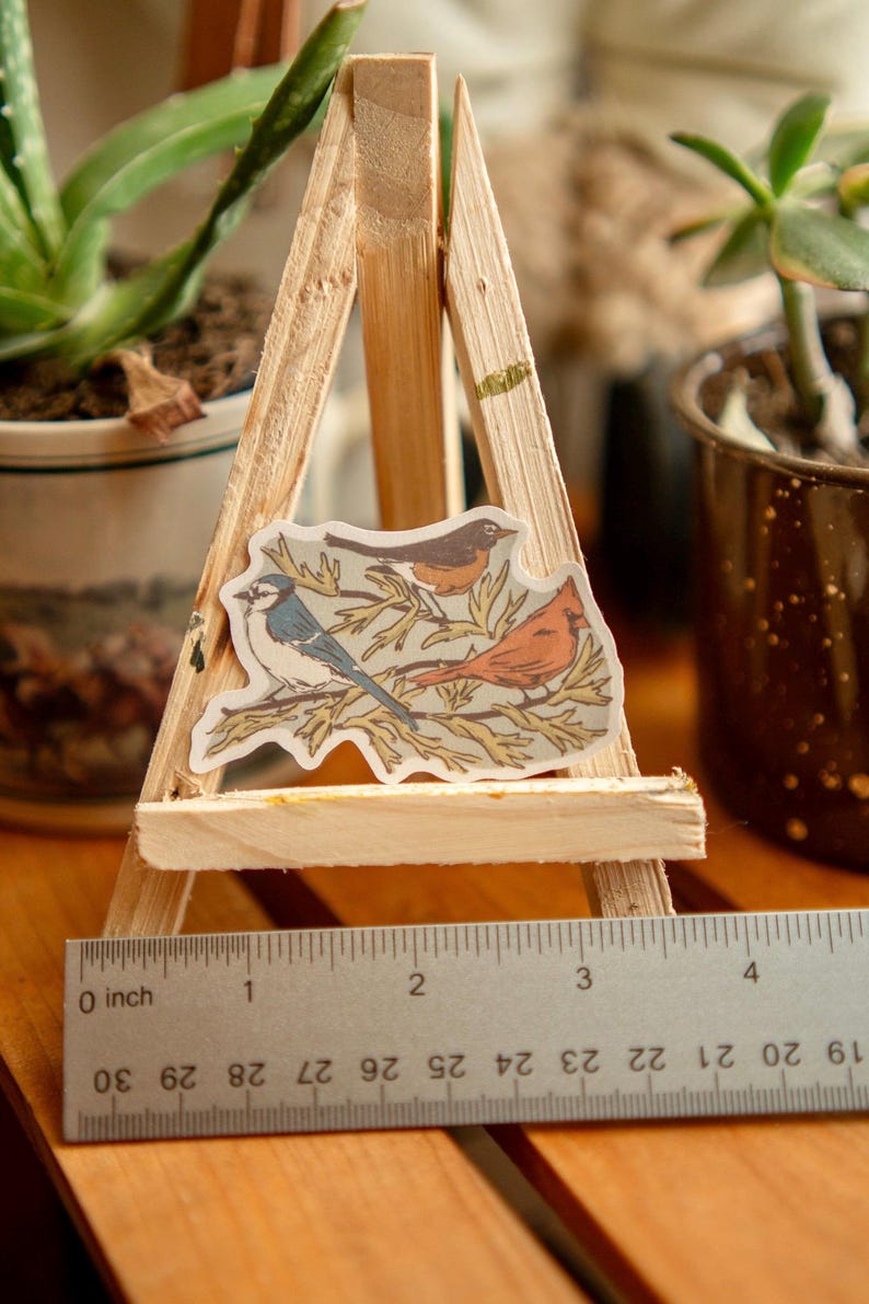Peut inclure: Un autocollant repr&eacute;sentant trois oiseaux perch&eacute;s sur des branches, expos&eacute; sur un petit chevalet en bois. Les oiseaux sont un geai bleu, un rouge-gorge et un cardinal. La largeur de l'autocollant est d'environ 7.6 cm.