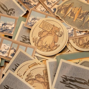 Op de afbeelding: Een verzameling diverse stickers met religieuze thema's. Ontwerpen omvatten berglandschappen, vissen en een beer met een bord met de tekst "Jezus wil je problemen horen - praat erover met hem". De stickers hebben een vintage esthetiek.