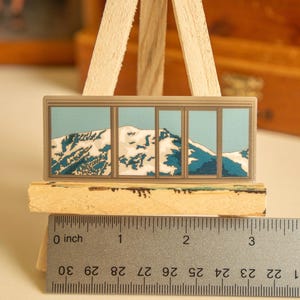 May include: A sticker of a mountain range with snow-capped peaks, viewed through a window with multiple panes. The sticker is approximately 2 inches wide and is displayed on a wooden easel.